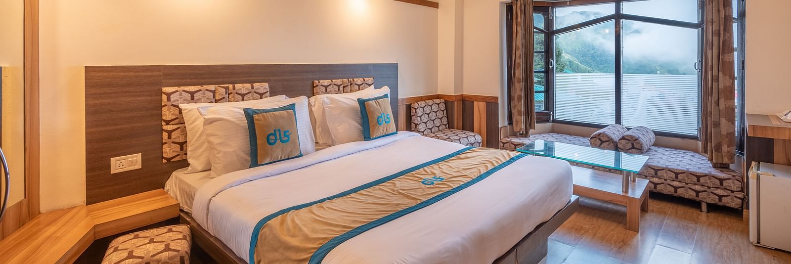 A hotel room with a large bed, a window overlooking a mountain view, and a seating area - DLS Meghavan Resort